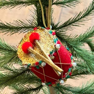 May include: A festive Christmas tree ornament shaped like a drum. The drum is red with gold sequins and trim, featuring red pom-poms and drumsticks. The ornament is suspended by a gold cord and is surrounded by green pine needles.