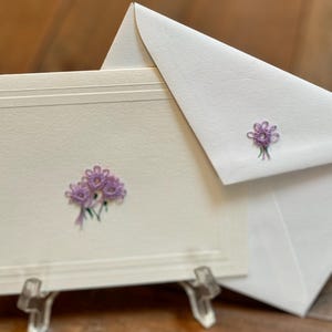 May include: Ivory stationery set featuring a card and envelope, both adorned with delicate purple floral embroidery. The card has a decorative border and is displayed on a clear acrylic stand. The envelope is tucked behind the card.