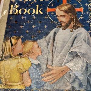 May include: A Catholic Child's Prayer Book with a blue cover featuring an illustration of Jesus with two children. The cover has gold lettering and a cross design. The image depicts a religious theme.