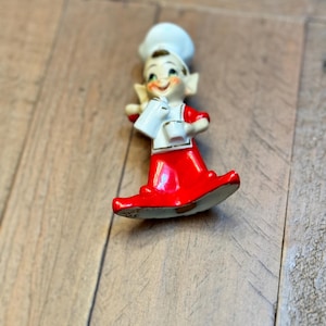 May include: A ceramic figurine of a smiling chef wearing a white toque and a red apron over a red outfit. The chef holds a white pitcher and a small white cup in their hands. The figure features painted facial details and pointed ears.