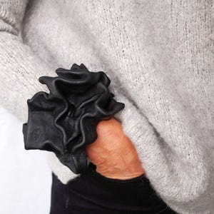 May include: Black leather cuff featuring a ruffled design. The cuff is worn over the sleeve of a light grey sweater. This statement cuff adds a touch of drama and sophistication to any outfit.