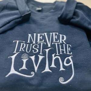 Pu&ograve; includere: Una felpa blu scuro con testo ricamato in bianco che recita "Never Trust the Living".