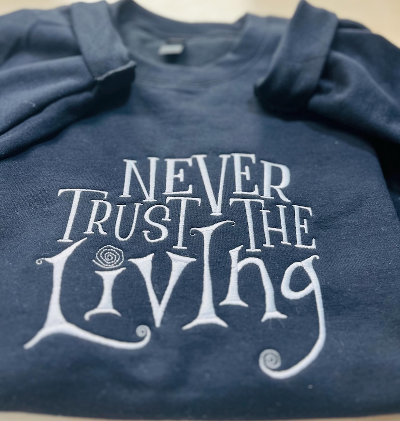 Pu&ograve; includere: Una felpa blu scuro con testo ricamato in bianco che recita "Never Trust the Living".