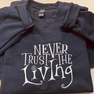 Pu&ograve; includere: Una felpa nera con il testo "Never Trust the Living" ricamato in bianco sul davanti.