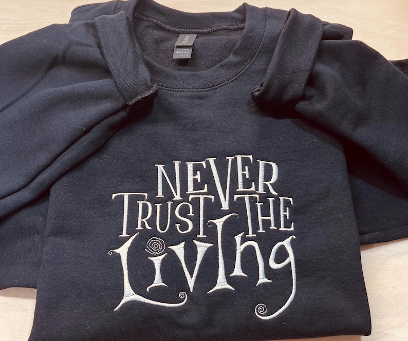 Pu&ograve; includere: Una felpa nera con il testo "Never Trust the Living" ricamato in bianco sul davanti.