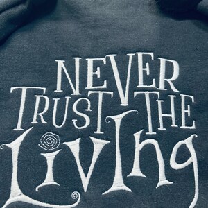Pu&ograve; includere: Una felpa nera con testo ricamato in bianco che recita "Never Trust the Living".