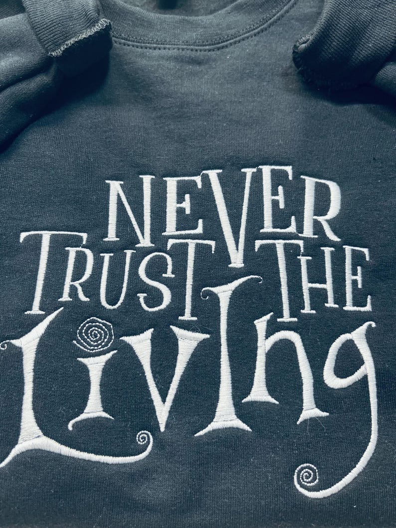 Pu&ograve; includere: Una felpa nera con testo ricamato in bianco che recita "Never Trust the Living".