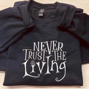 Pu&ograve; includere: Felpa nera con testo ricamato in bianco che recita "Never Trust the Living".