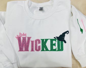 May include: White sweatshirt with a pink, green, and black embroidered design that says "WICKED". A crown is above the "W" and a witch's hat is above the "D".