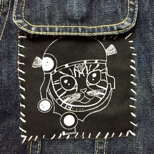 Peut inclure: Gros plan sur une poche de veste en jean bleu foncé avec un patch noir. Le patch représente un chat de dessin animé blanc portant un casque. Le patch est cousu à la poche avec du fil blanc. La veste a des coutures jaunes.
