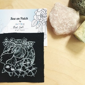 May include: Black fabric patch with a white outline of a bat with flowers and a full moon. The patch is 4" x 4" and says "Sew on Patch by Amy Tom Bat Cat 4" x 4" Patch".