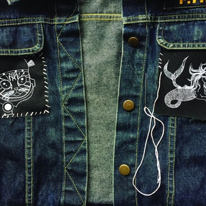May include: A blue denim jacket with two patches sewn on. The left patch features a black and white illustration of a cat wearing a helmet. The right patch features a black and white illustration of a mermaid.