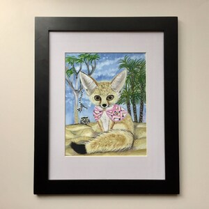 May include: A framed watercolor painting of a fennec fox wearing a pink and white patterned shirt. The fox is sitting in a desert landscape with palm trees and a blue sky.