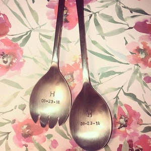 May include: Two silver serving spoons with long handles, one with tines. Each spoon is engraved with the letter "H" and the date "06-23-18". The spoons are set against a floral background with pink and red flowers.