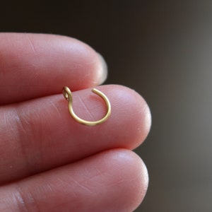 May include: A small, gold-coloured nose ring. The jewellery is a simple, circular design with a small loop at one end. The ring is held between two fingers, showcasing its delicate size and minimalist style.