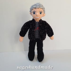 May include: A crocheted doll with gray hair, a black jacket, black pants, and a gray shirt. The doll has a friendly face with blue eyes and a smile.