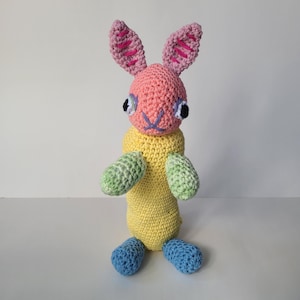 May include: A crocheted stuffed bunny toy with pink ears, yellow body, green arms, and blue feet.
