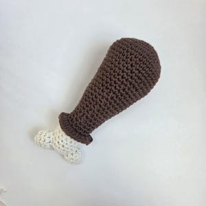May include: A handmade crochet toy shaped like a chicken leg. The toy features a brown drumstick and a white bone, with a dark brown ruffle at the base of the drumstick. The toy is made of soft yarn.
