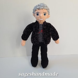 May include: A handmade crochet doll of a man with gray hair, glasses, and a smiling expression. He wears a black jacket over a gray shirt and black pants. The doll is standing on a white surface. The text "sageshandmade" is at the bottom.
