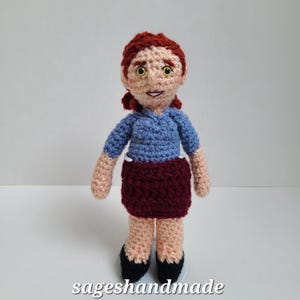May include: Crocheted doll with red hair, a blue shirt, a maroon skirt, and black shoes. The doll is standing on a white surface.