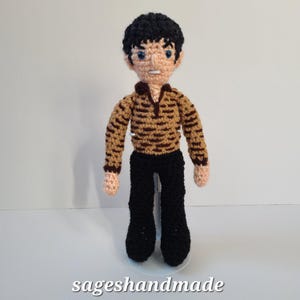 May include: Crocheted doll with black hair, brown and tan striped shirt, and black pants. The doll is standing on a white stand.