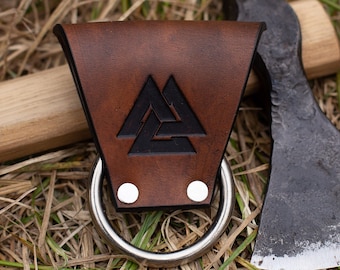 May include: A brown leather axe sheath with a metal ring and a black Valknut symbol. The sheath is attached to a wooden axe handle with a black metal axe head.