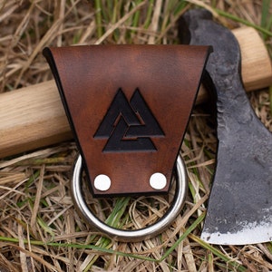 May include: A brown leather axe sheath with a metal ring and a black Valknut symbol. The sheath is attached to a wooden axe handle with a black metal axe head.