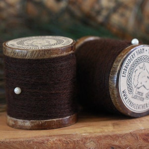 May include: Two spools of dark brown embroidery yarn with wooden sides and a paper label. The spools are on a wooden surface. One spool has a pearl pin. The label reads "Embroidery Yarn - 100% Wool - 54 meter".