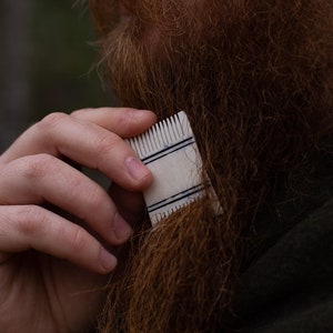 May include: A small, rectangular beard comb with black stripes is used to groom a long, reddish-brown beard. The comb is held in a person's hand, with the beard and a dark green garment in the background.
