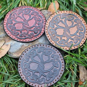 Puede incluir: Tres posavasos de cuero con un diseño de nudo celta. Los posavasos son de color marrón, beige y negro.