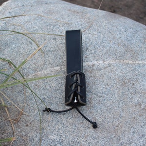 May include: A black leather strap with a grey leather lace-up closure. The strap is lying on a grey rock.