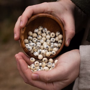 May include: A wooden bowl filled with small, round, off-white beads with black and blue circular designs. Some beads are spilling into the hands, which are holding the bowl. The bowl and beads are the focal point of the image.