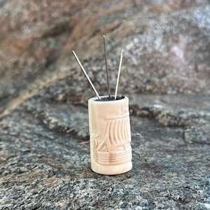 May include: A small, white, carved wooden container with a ship design on the side. Three needles are sticking out of the top of the container. The container is sitting on a gray rock.