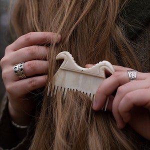 May include: A cream-colored comb with an ornate handle is used on long, light brown hair. The comb has a decorative, curved top and fine teeth. The person is wearing a silver ring with a geometric design.