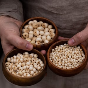 May include: Three wooden bowls filled with white wooden beads. The bowls are made of dark wood and are arranged in a row.