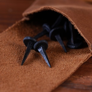 Pode incluir: Um close-up de uma bolsa de couro marrom com uma aba aberta, revelando uma coleção de tachas ou pregos de metal preto.