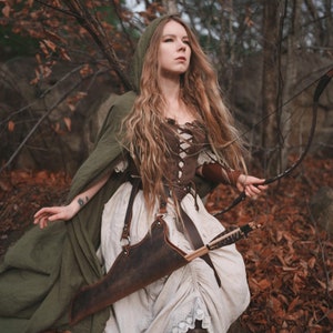 May include: A woman in a forest setting, wearing a brown leather corset, a white dress with lace trim, and a green hooded cloak. She is holding a bow and arrow and has a quiver of arrows strapped to her side.