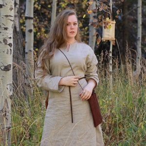 Puede incluir: Una mujer con un vestido largo de lino marr&oacute;n claro con un cintur&oacute;n de cuero marr&oacute;n y un bolso de cuero marr&oacute;n. Est&aacute; de pie en un bosque con &aacute;rboles altos y hierba verde.