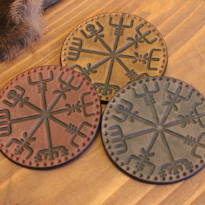 May include: Three round leather coasters with a brown, dark brown, and black color scheme. Each coaster features a circular design with a compass rose and runes.