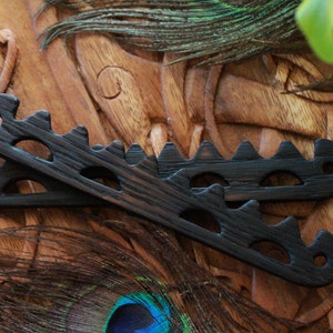 May include: Two dark wooden hair combs with a unique, openwork design featuring a series of peaks and rounded cutouts. The combs are displayed on a carved wooden surface, with peacock feathers adding a decorative touch.