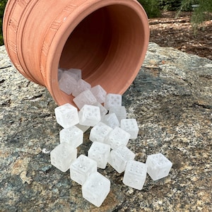 May include: A terracotta-colored ceramic pot tipped over, spilling clear, cube-shaped dice onto a textured rock surface. The dice have small, indented dots representing numbers. The pot has a ridged design around its rim.