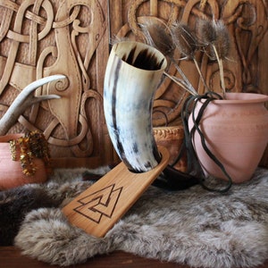 May include: A Viking drinking horn made of natural horn, resting on a wooden stand with a carved symbol. The horn has a light and dark brown color pattern. The stand is made of light-colored wood. The background features carved wooden panels and a fur rug.