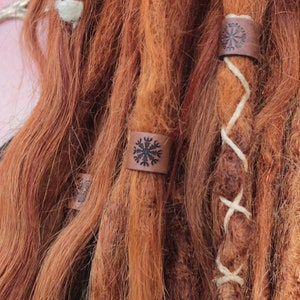 May include: A woman with long, red dreadlocks. Her hair is decorated with leather bands with a snowflake design. She is wearing a silver necklace with a pendant in the shape of antlers.