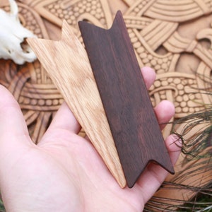 Op de afbeelding: Twee houten pijlpunten, een lichtbruin en een donkerbruin, worden in een hand gehouden. De pijlpunten zijn puntig en hebben een driehoekige vorm.