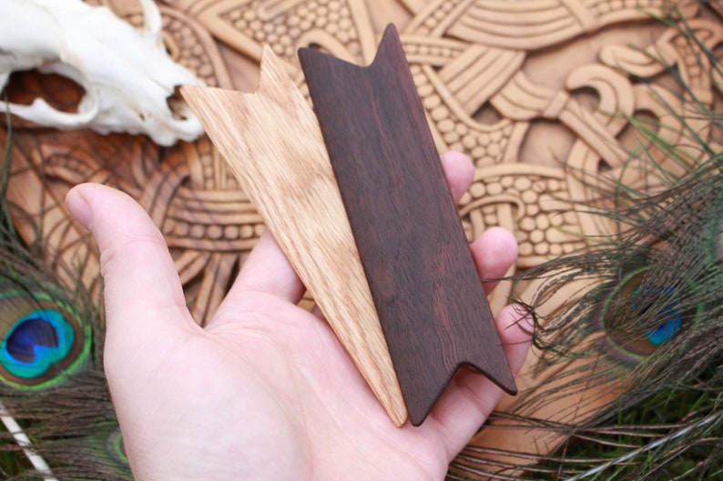 Op de afbeelding: Twee houten pijlpunten, een lichtbruin en een donkerbruin, worden in een hand gehouden. De pijlpunten zijn puntig en hebben een driehoekige vorm.