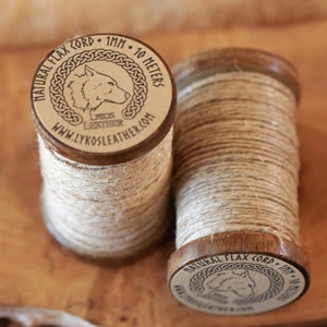 May include: Two spools of natural flax cord, each wound with beige twine and topped with a wooden cap. The caps are engraved with a wolf head logo and the text "NATURAL FLAX CORD - 1MM - 10 METERS" and "WWW.LYKOSLEATHER.COM".