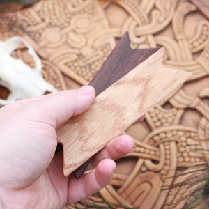 Op de afbeelding: Een hand houdt twee houten pijlpunten vast, een lichtbruin en een donkerbruin, tegen een achtergrond van een gegraveerd houten paneel met een Keltisch knoopontwerp en pauwenveren.