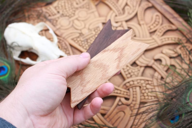 Op de afbeelding: Een hand houdt twee houten pijlpunten vast, een lichtbruin en een donkerbruin, tegen een achtergrond van een gegraveerd houten paneel met een Keltisch knoopontwerp en pauwenveren.