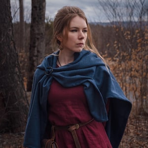 May include: A woman wearing a long, burgundy dress with a blue shawl and a brown leather belt. She is standing in a forest setting with trees and fallen leaves.