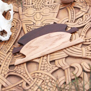 May include: Two wooden hair combs, one dark brown and one light brown, rest on a carved wooden surface with intricate patterns. A skull is visible in the upper left corner. The combs are designed with a unique shape, suggesting a Viking or Norse theme.